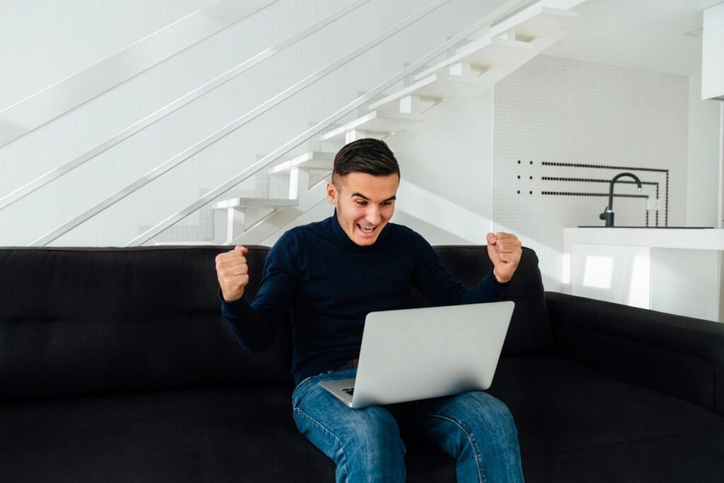 A photo of a New Hampshire man double fist pumping in the air because he just filed bankruptcy from his couch!  The laptop on his lap is neutral, he wears blue jeans and a black sweater, sits on top a black couch in front of a white staircase.  